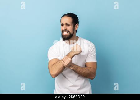 Porträt eines kranken Mannes mit Bart in weißem T-Shirt, der mit Schmerzen steht, schmerzende Handgelenke massiert, Handverletzungen oder Verstauchungen leidet. Innenaufnahme des Studios isoliert auf blauem Hintergrund. Stockfoto