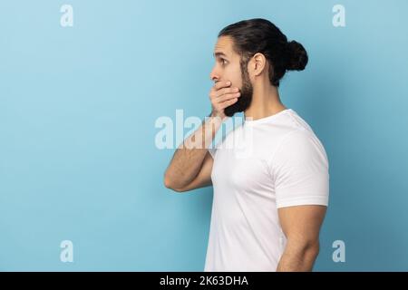 Seitenansicht eines Mannes mit Bart und weißem T-Shirt, der den Mund bedeckt, möchte keine Gerüchte verbreiten, schockiert, als er plötzliche Nachrichten vom Gesprächspartner hörte. Innenaufnahme des Studios isoliert auf blauem Hintergrund. Stockfoto