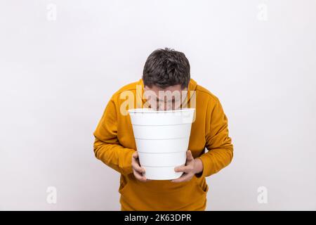 Porträt eines ungesunden Mannes mittleren Alters, der plötzliches Erbrechen, Übelkeit, Mülltonne in den Händen hält und einen Kapuzenpullover im urbanen Stil trägt. Innenaufnahme des Studios isoliert auf weißem Hintergrund. Stockfoto