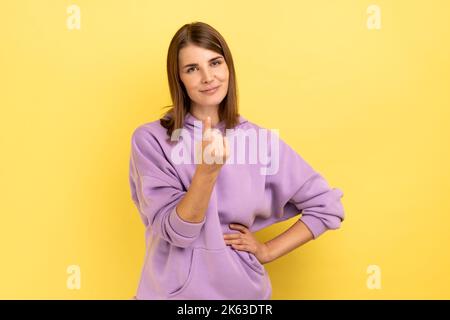 Kommen Sie hierher. Porträt einer attraktiven Frau, die eine winkende Geste macht, die zum Kommen einlädt, flirtet, die Kamera anschaut und einen purpurnen Hoodie trägt. Innenaufnahme des Studios isoliert auf gelbem Hintergrund. Stockfoto