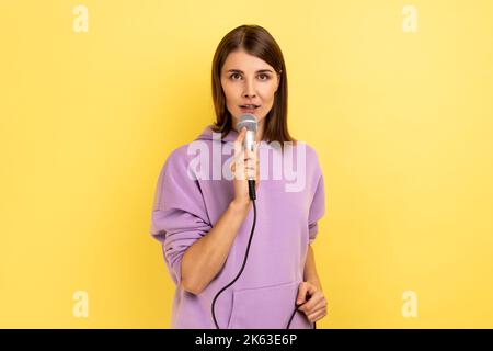 Junge Erwachsene schöne Frau Reporterin hält Mikrofon, berichtet und sagt Meinung, diskutiert wichtige Themen, trägt lila Hoodie. Innenaufnahme des Studios isoliert auf gelbem Hintergrund. Stockfoto