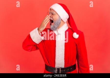 Älterer Mann mit grauem Bart, der das weihnachtsmann-Kostüm trägt und den Mund mit dem Arm bedeckt, fühlt sich nach einer schlaflosen Nacht schläfrig, Mangel an Energie. Innenaufnahme des Studios isoliert auf rotem Hintergrund. Stockfoto