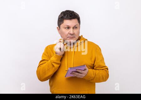 Nachdenklicher Mann, der mit nachdenklicher Ausdrucksweise steht und ein Notizbuch in der Hand hält, über Geschäftsidee nachdenkt, über Zukunftspläne nachdenkt und einen Hoodie im urbanen Stil trägt. Innenaufnahme des Studios isoliert auf weißem Hintergrund. Stockfoto