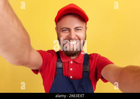 Fröhlicher, aufgeregt Arbeiter mit roter Mütze und blauer Uniform, der Selfie macht, die Kamera mit einem toothy Lächeln aus der Perspektive des Fotos anschaut. Innenaufnahme des Studios isoliert auf gelbem Hintergrund. Stockfoto