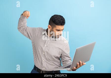 Wütender Geschäftsmann, der mit einem Notebook-Bildschirm zuschlägt, mit tollem Ausdruck aussieht, beim Online-Gespräch mit einem Boxdrohen zu schlagen droht und ein gestreiftes Hemd trägt. Innenaufnahme des Studios isoliert auf blauem Hintergrund Stockfoto