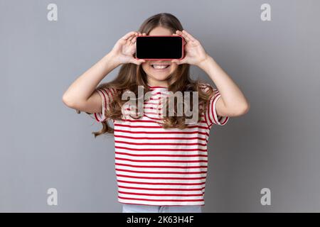 Porträt eines kleinen Mädchens mit gestreiftem T-Shirt, das die Augen mit dem Handy bedeckt und lächelt, unbekanntes Kind, das sich mit dem Handy versteckt, anonymer Benutzer. Innenaufnahme des Studios isoliert auf grauem Hintergrund. Stockfoto