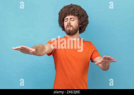 Porträt eines Mannes mit Afro-Frisur, der sich mit geschlossenen Augen bewegt, den Raum mit Händen berührt, mit Dunkelheit verwirrt, Sehstörungen, Sehstörungen. Innenaufnahme des Studios isoliert auf blauem Hintergrund. Stockfoto