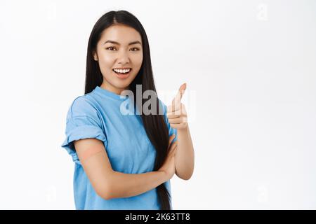 Das enthusiastische asiatische Mädchen wurde geimpft, hat nach dem Covid-Impfstoff ein Pflaster auf der Schulter, lächelt und zeigt auf das Transparent des Kopierraums, das zu sehen ist Stockfoto