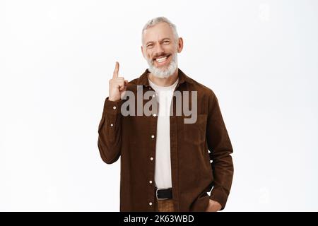 Bild eines lächelnden, hübschen bärtigen Mannes mit grauen Haaren, eines älteren Mannes, der mit dem Finger nach oben zeigt, Werbung zeigt und auf weißem Hintergrund steht Stockfoto