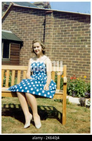 Eine hübsche junge Dame in einem blau-weißen Polka-Dot-Kleid, die am Ende einer Bank in einem gut-gruseligen vorstädtischen britischen Garten sitzt - September 1959. Stockfoto