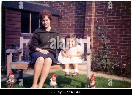 Eine junge Frau, die in einem gepflegten Vorstadtgarten auf einer Holzbank sitzt, zusammen mit einem jungen Verwandten (möglicherweise ihrer Nichte?) - Sie sind von Gartengnomen umgeben! Juli 1961 Stockfoto