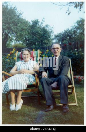 Ein fröhliches älteres Paar, das in leichten ausklappbaren Gartenstühlen aus Holz und Leinwand in einem gepflegten Vorstadtgarten sitzt - September 1959 Stockfoto