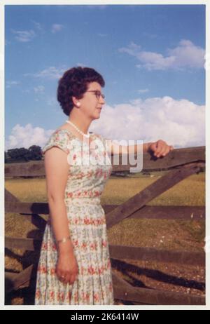 Eine elegant gekleidete Frau mittleren Alters in einem floral gemusterten Sommerrock und Perlen, die ziemlich wehmütig in die Ferne blickt, während sie sich an einem Tor lehnt, das in ein Landfeld führt - August 1962 Stockfoto