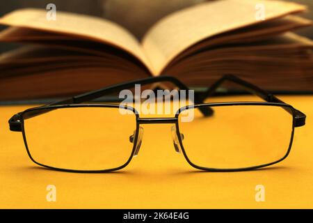 Brille in schwarzem Rahmen auf gelbem Hintergrund. Im Hintergrund ist ein offenes Buch. Welttag des Sehens. Stockfoto