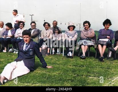 Eine Gruppe von Erwachsenen, die hintereinander sitzen, mit einem kleinen Mädchen in der Mitte, und einem männlichen Redcoat, der auf dem Gras im Vordergrund sitzt, in Butlins Ferienlager, Filey, North Yorkshire. (1 von 2) Stockfoto