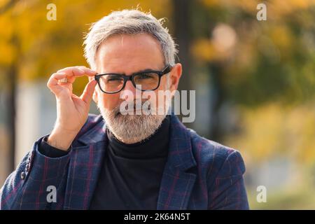 Brillen kommerzielles Konzept. Nahaufnahme eines hübschen eleganten kaukasischen Mannes im Alter von 60s Jahren im Freien mit grauen Gesichtshaaren, die seine Brille mit schwarzem Rahmen mit einer Hand berührten. Warme Herbstsonne scheint auf einem Teil seines Gesichts. Unscharfer Hintergrund. Hochwertige Fotos Stockfoto