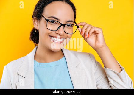 Nahaufnahme einer wunderschönen erfolgreichen Latino- oder brasilianerin mit Brille, stilvoll gekleidet, hr-Manager, Immobilienmakler, Makler, Blickt auf die Kamera, lächelt, steht auf einem isolierten orangefarbenen Hintergrund Stockfoto