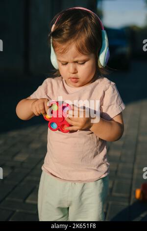 Kleines Mädchen steht und hält ein Spielzeug in den Händen, trägt Ohrenstöpsel und ist abends kühl. Baby und sonniges Wetter Stockfoto