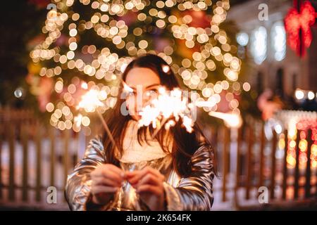 Junge glückliche Frau mit Wunderkerzen, während sie draußen in der Stadt gegen den beleuchteten Weihnachtsbaum steht Stockfoto