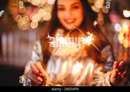 Junge glückliche Frau mit Wunderkerzen, während sie draußen in der Stadt gegen den beleuchteten Weihnachtsbaum steht Stockfoto