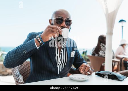 Ein Porträt eines kahlen Afrikaners in Sonnenbrille und eines modischen Sommerkanzers, der in einem Straßencafé sitzt und Kaffee trinkt, mit einem selektiven FOC Stockfoto