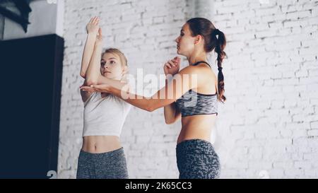 Der professionelle Yogalehrer unterrichtet neue Schüler, während er im leichten Wellness-Studio individuelle Übungen macht, um ihre Arme zu drehen. Mädchen lachen während der Übungen, entspannende Atmosphäre. Stockfoto