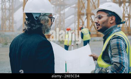 Vermesser arbeiten mit Geräten, während der Architekt das Projekt mit dem Vorarbeiter diskutiert, der Pläne auf der Baustelle betrachtet Stockfoto