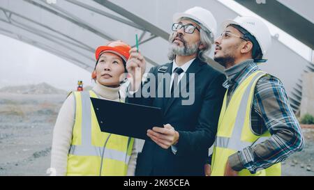 Ingenieure Asiatin und arabischer Mann sprechen mit Projektmanagerin, die auf der Baustelle zusammen steht und sich umschaut und Papiere überprüft Stockfoto