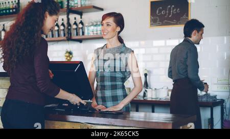 Höfliche lächelnde Angestellte des netten modernen Cafés verkaufen Kaffeegetränke zum Mitnehmen, während zufriedene Kunden mit dem Smartphone bezahlen. E-Geld- und Food-Service-Konzept. Stockfoto