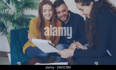 Liebenswert Paar überprüft Papiere während des Treffens mit makler in schöne neue Wohnung. Junge Menschen sitzen auf dem Sofa, umarmen sich und unterhalten sich mit dem Makler. Stockfoto