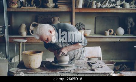 Qualifizierte ältere männliche Töpfer produziert schöne Keramik Topf auf Drehrad am Arbeitsplatz. Entstehungsprozess, traditionelle Keramik und Interessantes Hobby-Konzept. Stockfoto