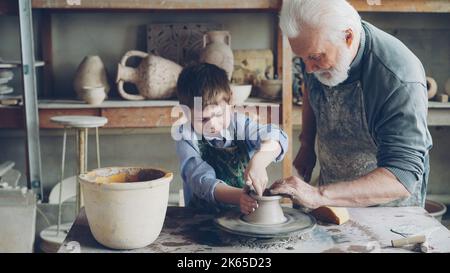 Konzentrierter kleiner Junge lernt mit Ton auf professionellem Wurfrad im Töpferunterricht in traditioneller Werkstatt zu arbeiten. Sein erfahrener Lehrer hilft ihm. Stockfoto