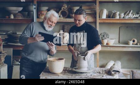 Der junge männliche Bildhauer macht eine Tonfigur auf dem Wurfrad, während sein Vater ihm eine Tablette zeigt, Männer diskutieren über Töpferwaren und schauen sich den Bildschirm an. Traditionelle Besetzung und modernes Technologiekonzept Stockfoto