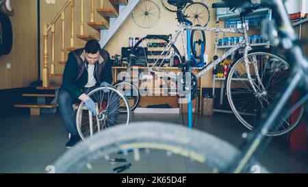 Hübscher männlicher Mechaniker fixiert das Hinterrad mit einem Schraubenschlüssel, während er am modernen Arbeitsplatz auf einem Holzhocker sitzt. Zyklen, Ersatzteile, Werkzeuge und Geräte sind sichtbar. Stockfoto