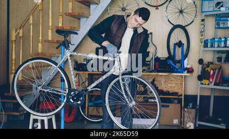 Ein junger erfahrener Wartungsmann montiert das Fahrrad, stellt das Lenkrad auf und fixiert es, während er an seinem Arbeitsplatz mit Ohrhörern Musik hört. Reparatur- und Personalkonzept. Stockfoto