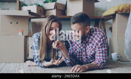 Fröhlicher bärtiger Kerl gibt Schlüssel zu seiner Freundin, die auf dem Boden der neuen Wohnung liegt. Zuneigung, junge Menschen, romantische Beziehung und Wohnkonzept. Stockfoto