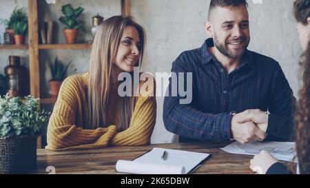 Attraktive Maklerin verkauft Haus an junges Paar, Menschen unterzeichnen Dokumente und schütteln die Hände, makler gibt Schlüssel zu neuen Besitzer, er küsst seine Frau. Stockfoto