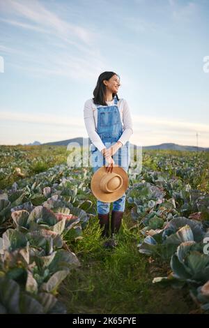 Bäuerin, die auf einem Kohlfeld auf einem Bauernhof steht. Junge Brünette Weibchen mit einem Strohhut und Gummistiefeln, die über ein Feld von Bio-Gemüse schauen Stockfoto