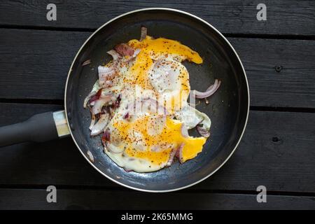 Gebratenes Hühnerei mit Zwiebeln in einer Pfanne steht auf einem schwarzen Holztisch, Rührei zum Frühstück Stockfoto