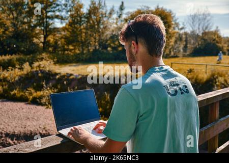 Ein junger Mann, der im Sommer mit einem Laptop am Geländer einer Brücke arbeitet Stockfoto