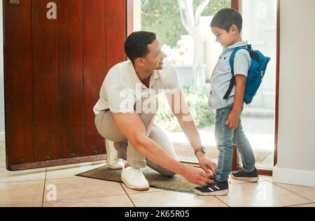 Glücklicher alleinerziehender Vater, der seine Söhne morgens vor der Schule mit Schnürsenkeln schnürt. Liebenswert gemischt Rennen kleinen Jungen mit einem Rucksack und immer Hilfe von Stockfoto