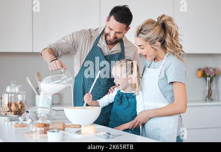 Vater gießt einen Krug Milch in den Teig. Glücklicher Vater beim Backen mit seiner Tochter. Kaukasische Familie backt zusammen.lächelnde Eltern helfen ihrem Kind Stockfoto