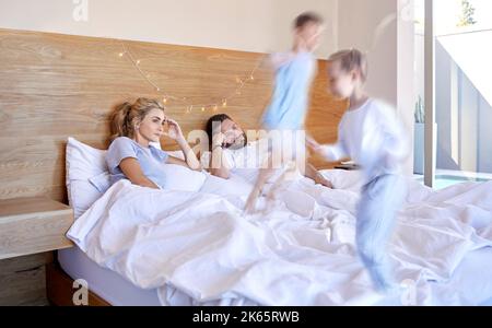 Müde und gestresste Eltern, die unter Kopfschmerzen leiden, während adhd-Kinder zu Hause auf das Bett springen. Laute, freche Geschwister ärgern ihre Mutter und ihren Vater Stockfoto