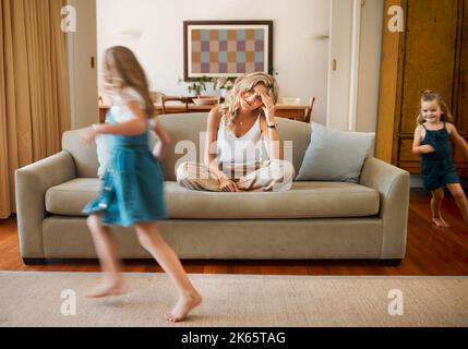 Junge unglückliche kaukasische Mutter, die unter Kopfschmerzen leidet, während ihre Töchter zu Hause in der Lounge herumlaufen und spielen. Kleine Geschwister sind verspielt Stockfoto