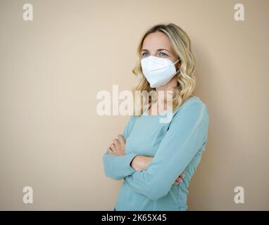 Hübsche junge Frau mit lockigen blonden Haaren, die FFP2 Schutzmaske vor braunem Hintergrund im Studio trägt Stockfoto