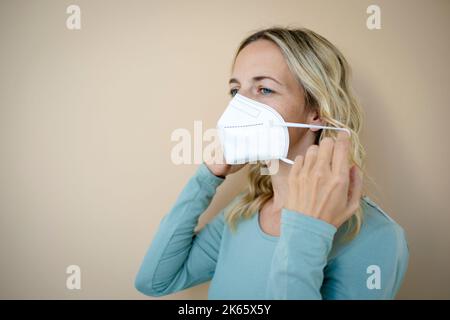 Hübsche junge Frau mit lockigen blonden Haaren, die FFP2 Schutzmaske vor braunem Hintergrund im Studio trägt Stockfoto