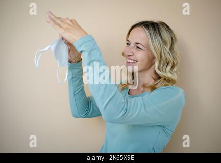 Hübsche junge Frau mit lockigen blonden Haaren, die FFP2 Schutzmaske vor braunem Hintergrund im Studio trägt Stockfoto