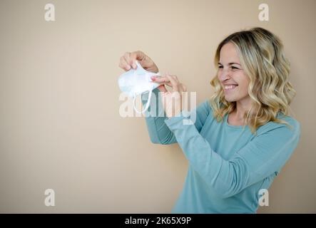 Hübsche junge Frau mit lockigen blonden Haaren, die FFP2 Schutzmaske vor braunem Hintergrund im Studio trägt Stockfoto