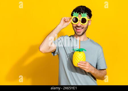 Foto von Millennial funky bärtigen Brunet Kerl aufgeregt gute Laune halten Brillen Sonnenbrillen trinken frischen Saft Sommer isoliert auf gelbem Hintergrund Stockfoto