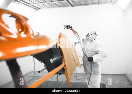 Mann in Schutzmaske und Kleidung sprüht Lack auf die Motorhaube mit einer Spritzpistole in einer Lackierkabine. Stockfoto